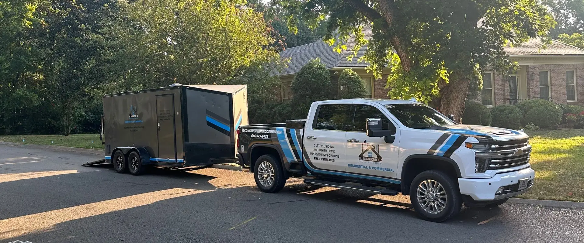 A white and blue J.L. Construction LLC Chevrolet service truck parked on a residential street, towing a large black enclosed equipment trailer with matching company branding.
