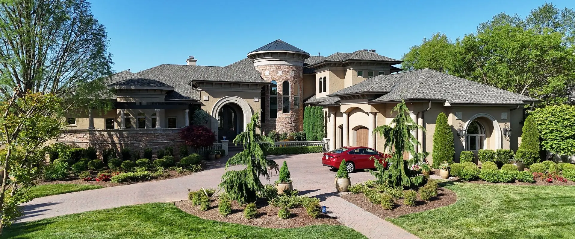 A wide-angle shot of a sprawling, multi-story luxury estate featuring Mediterranean-style architecture with tan stucco walls, stone accents, a circular turret, and meticulously manicured green landscaping.