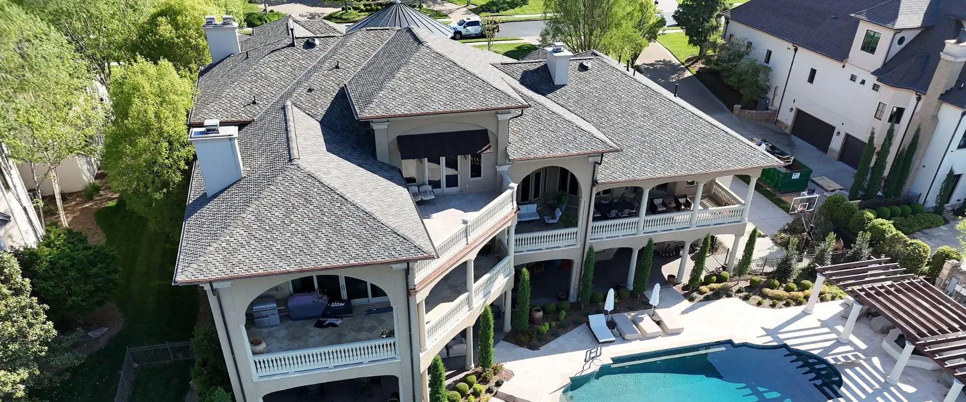 A high-angle drone shot looking down at the rear of a multi-story Mediterranean-style home, showcasing its expansive newly shingled roof, tiered stone balconies, and an adjacent luxury pool area with lounge seating.