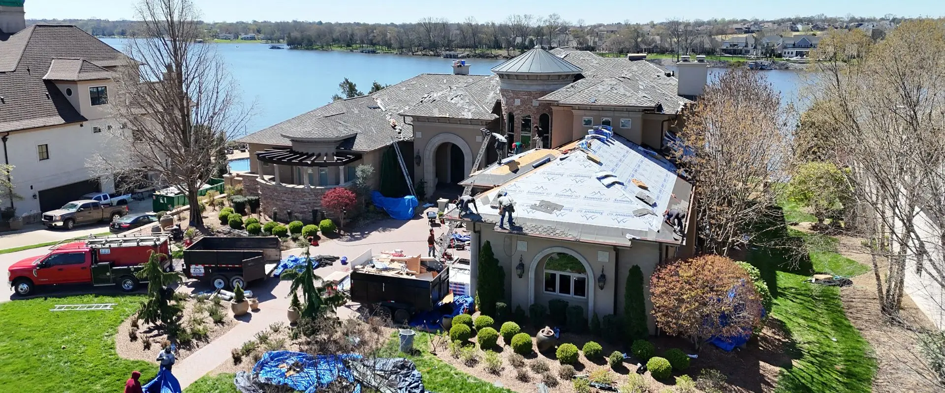 An elevated view of a luxury lakefront estate undergoing a roof replacement, with workers installing blue synthetic underlayment on the garage roof while a red service truck and dumpsters are parked in the driveway.