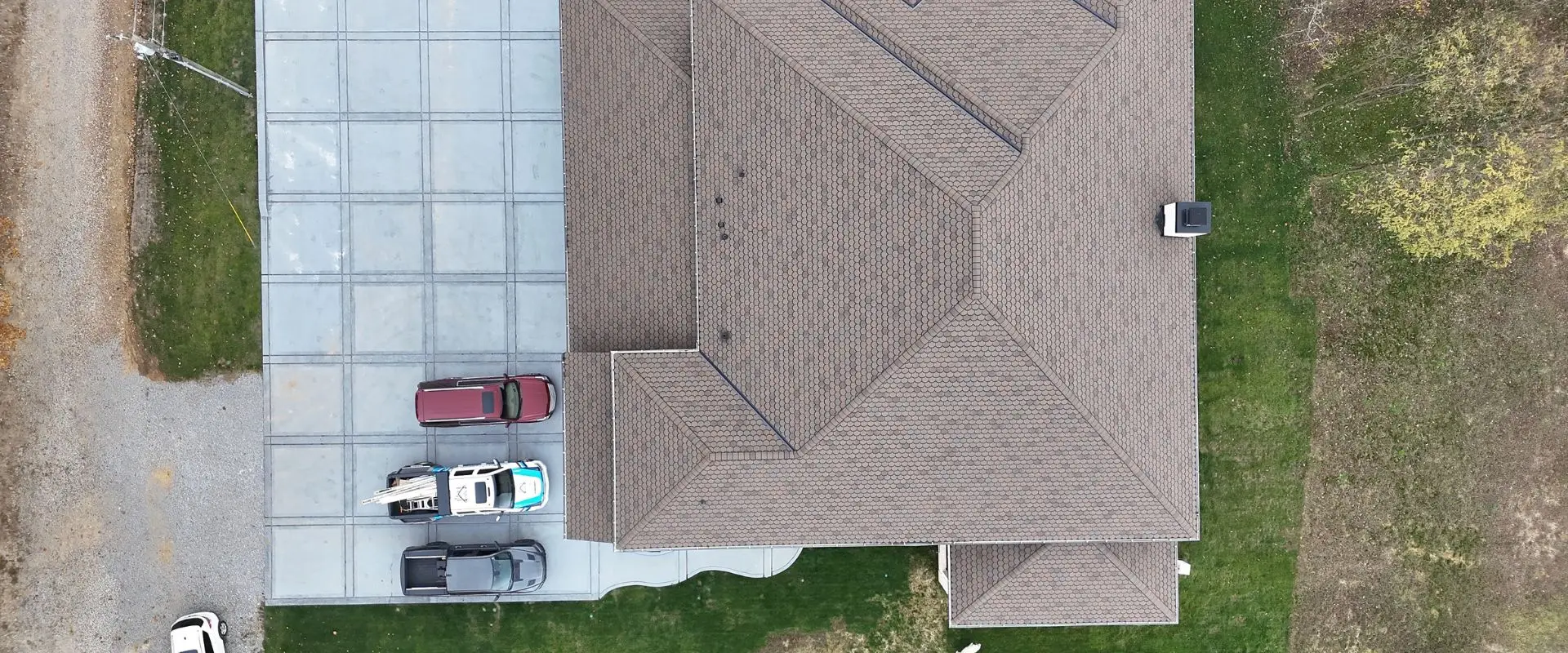 An overhead aerial view of a large white brick residence featuring a newly installed brown scalloped architectural shingle roof. The property includes a large, square-tiled gray concrete driveway where a maroon SUV and two branded company service trucks are parked.