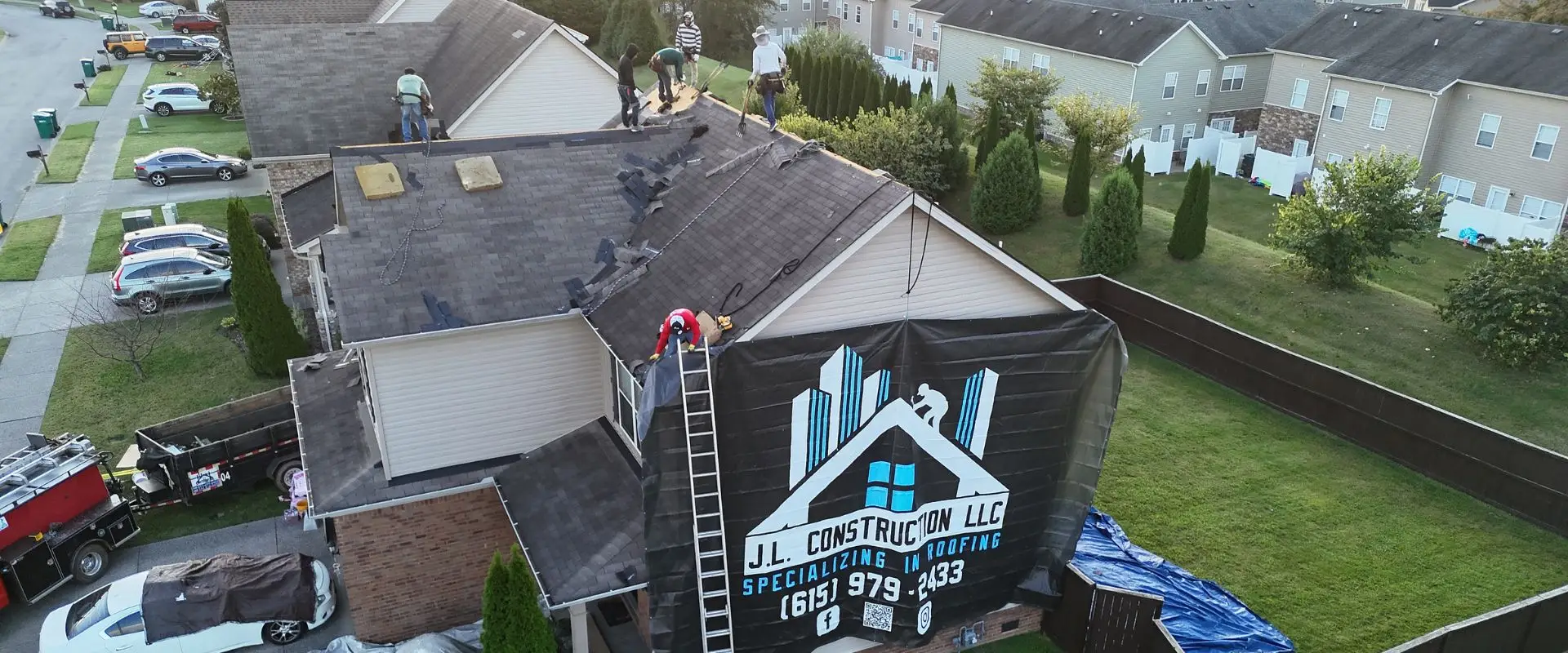 An aerial view of a roofing crew tearing off shingles from a residential home, with a large black J.L. Construction LLC banner hanging from the side of the house and blue tarps protecting the lawn.