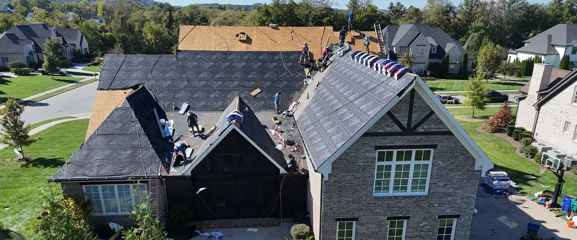 An aerial shot of a residential roofing crew active on a large brick home, with workers installing synthetic black underlayment over plywood decking and lining up colorful bundles of shingles along the roof's peaks.