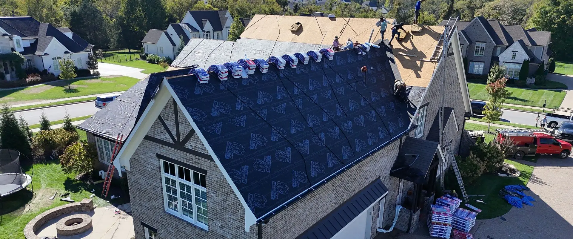 An aerial view of a roofing team installing black synthetic underlayment on the gabled roof of a brick home, with rows of blue shingle bundles lined up along the ridge and workers on ladders and the roof deck.