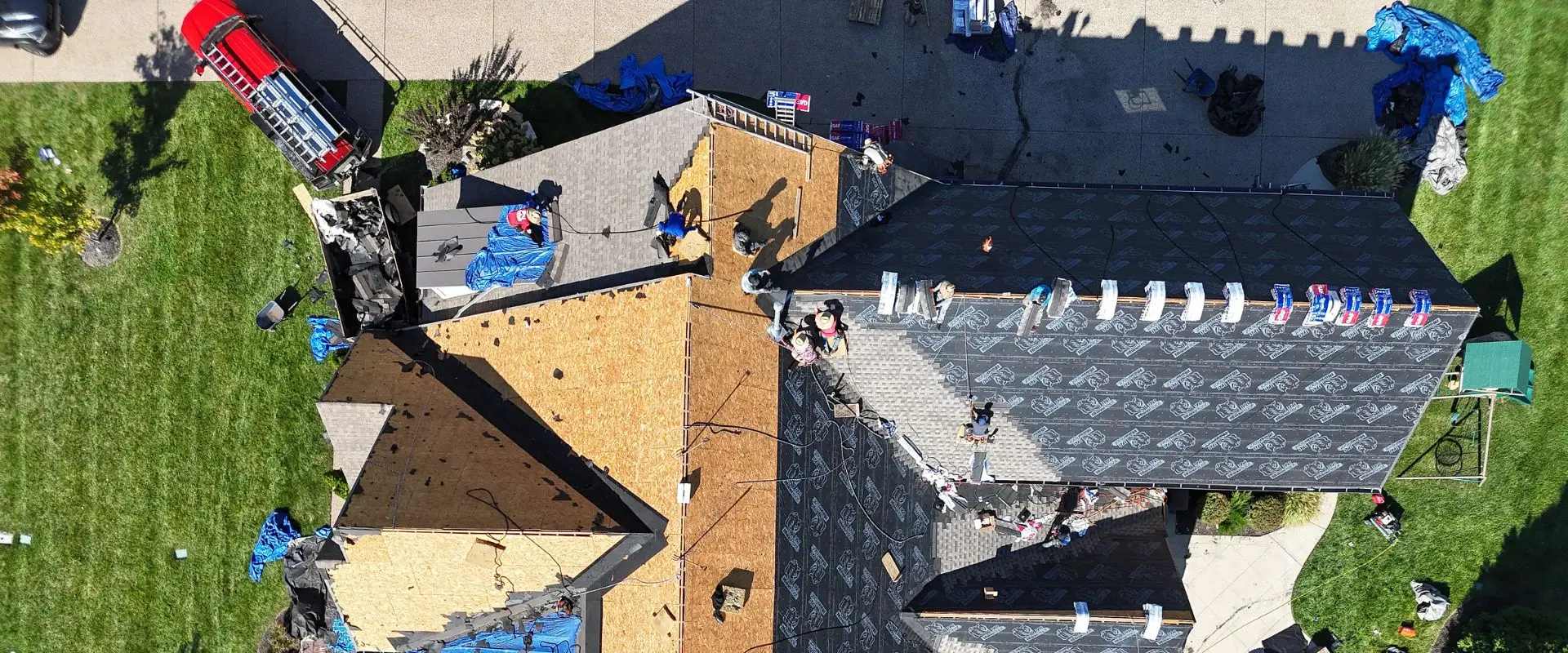 An aerial top-down view shows a roofing crew actively installing new shingles on a large residential home. Workers are distributed across the roof, laying down synthetic underlayment over plywood decking and securing new shingles, while a red service truck and a debris dumpster are parked on the driveway below.