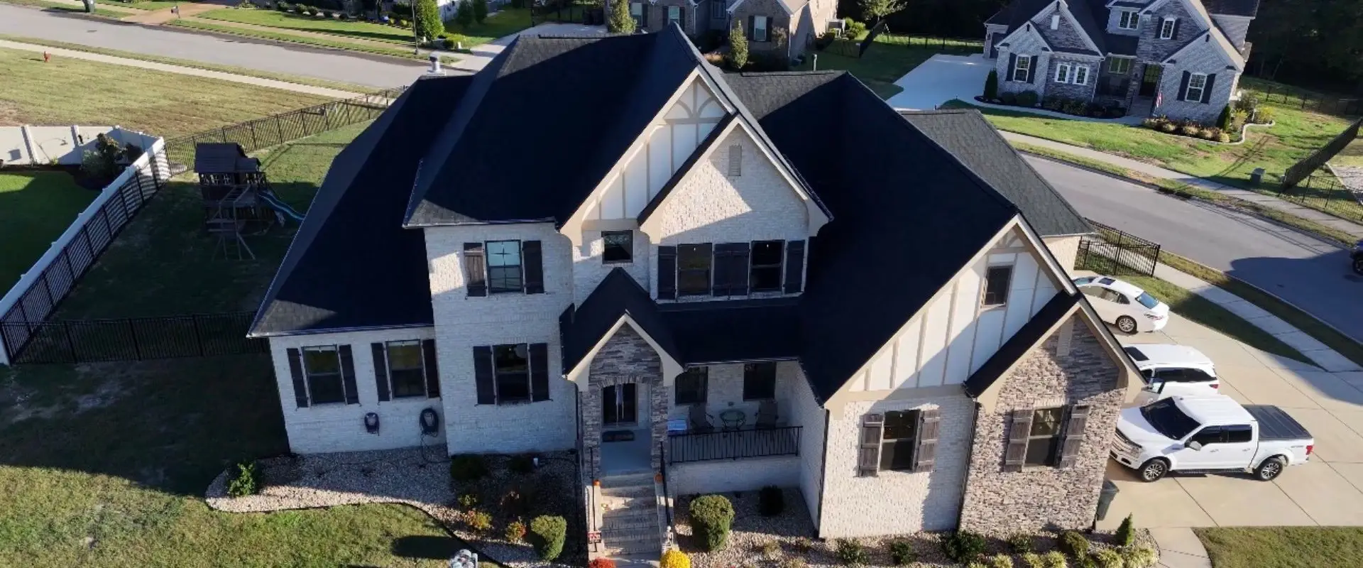 An aerial high-angle view of a large, two-story luxury home featuring a newly installed dark shingle roof, light stone and brick siding, and dark shutters, with a white pickup truck parked in the driveway.