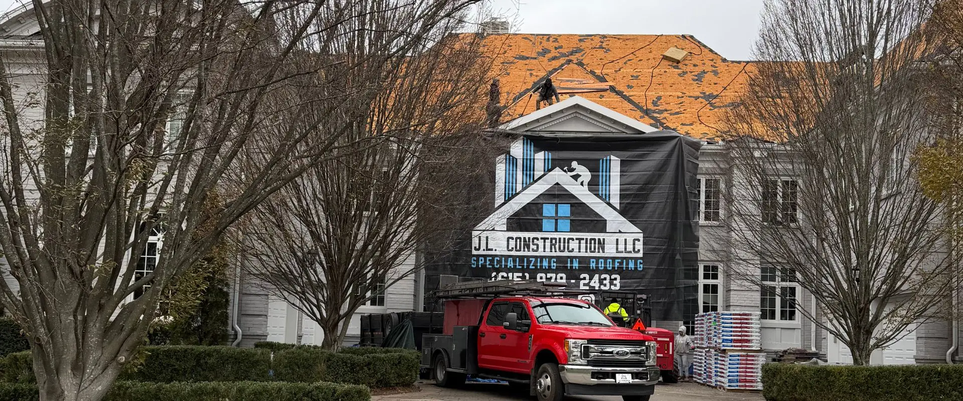 A red Ford service truck from J.L. Construction LLC parked in front of a gray stone mansion where roofers are installing a new roof, featuring stacks of shingles in the foreground and a large black company banner on the facade.