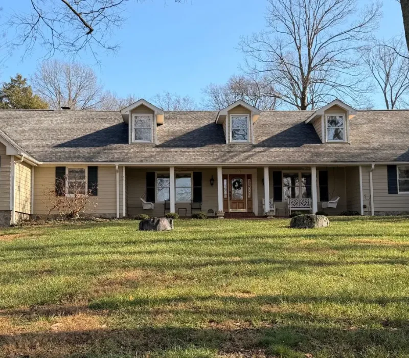A wide-angle front view of a long, single-story ranch-style home with beige siding, dark shutters, and three prominent dormer windows set against a grassy lawn and mature trees.