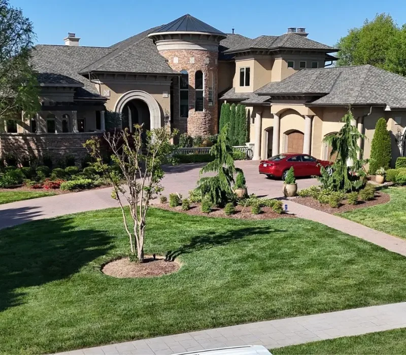 An aerial view of a luxury Mediterranean-style estate featuring a sprawling lawn, mature trees, and a brick driveway leading to a multi-car garage, situated along the edge of a calm lake.