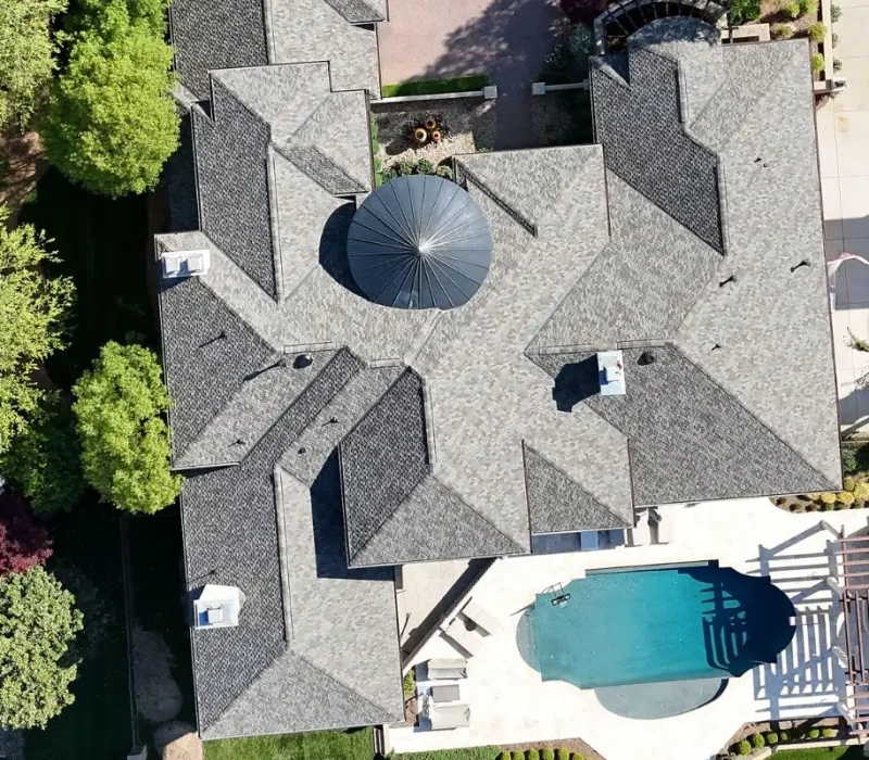 An aerial top-down view of a complex gray asphalt shingle roof on a luxury estate, featuring a prominent dark metal conical turret roof and an adjacent backyard pool area.