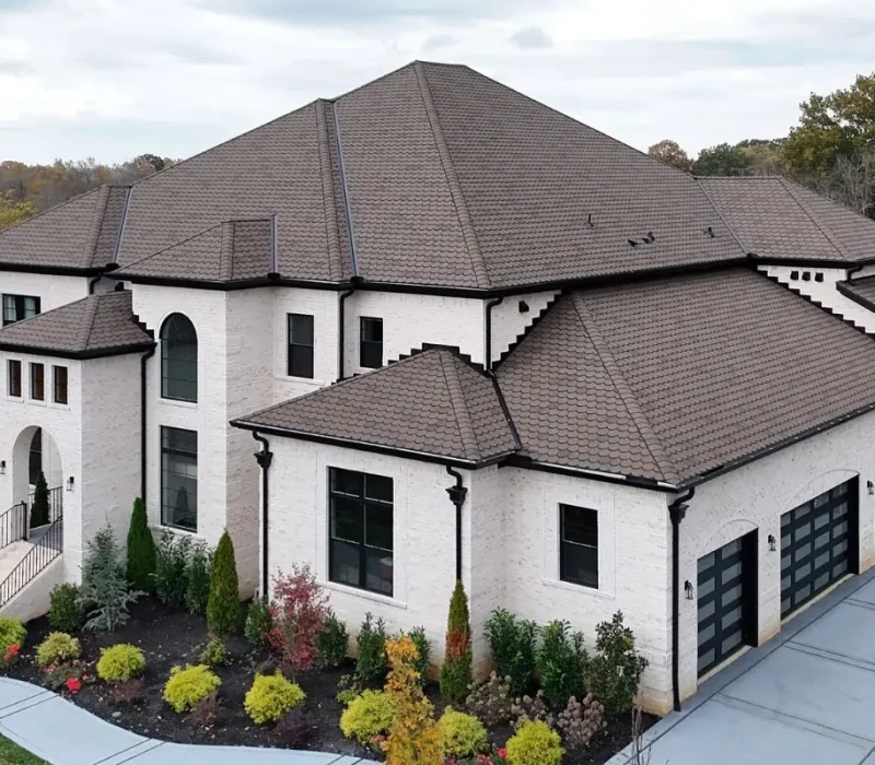 An aerial view of a large, two-story white brick home featuring a newly installed brown shingle roof with multiple gables and black gutters, alongside a spacious concrete driveway where a maroon SUV and service trucks are parked.