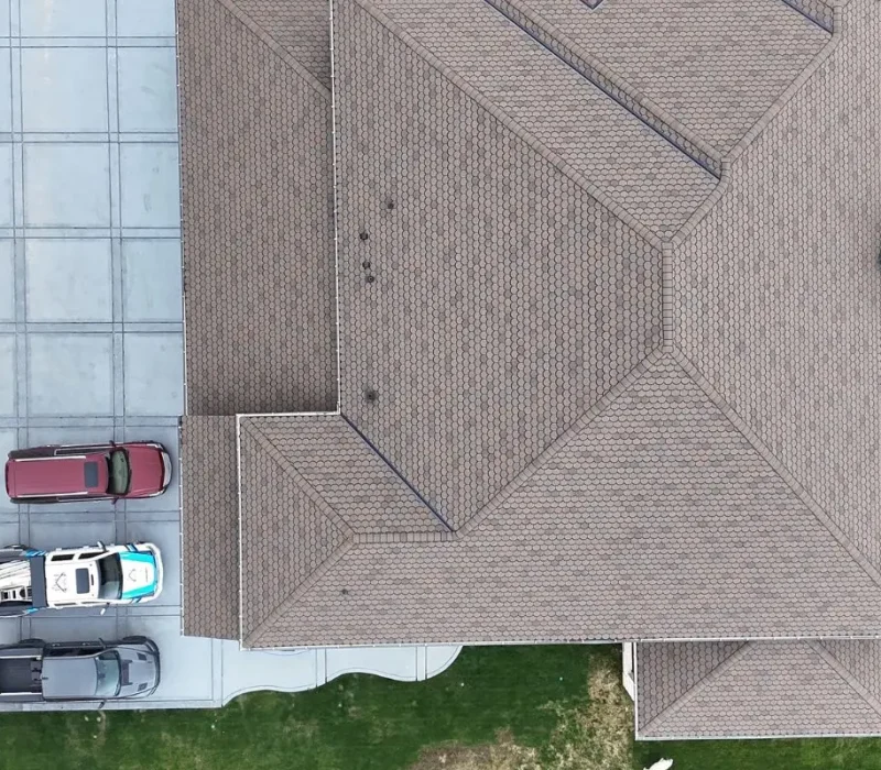 An overhead aerial view of a large white brick residence featuring a newly installed brown scalloped architectural shingle roof. The property includes a large, square-tiled gray concrete driveway where a maroon SUV and two branded company service trucks are parked.