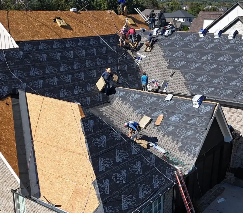An aerial view of a brick house mid-renovation, with roofing contractors installing black synthetic underlayment over the plywood decking and bundles of shingles lined up along the roof ridge.