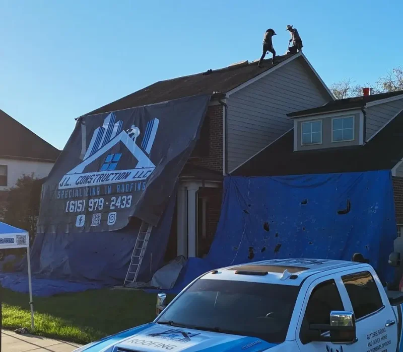 An ground-level view of a residential roofing project site featuring a white company truck in the foreground and a red service truck in the driveway. The two-story home is partially covered by large blue tarps and a black J.L. Construction LLC banner, while a company-branded pop-up tent is set up on the front lawn.
