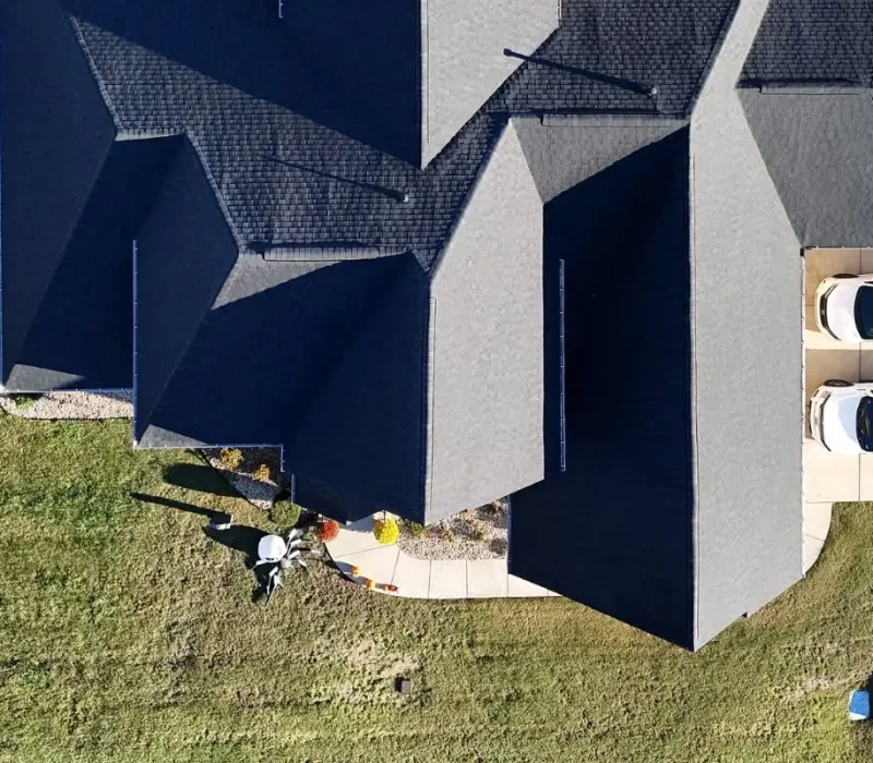 An aerial top-down view of a large residential property featuring a newly installed dark charcoal shingle roof with multiple complex gables. Three white vehicles are parked on the large concrete driveway adjacent to the house, and a manicured lawn surrounds a stone walkway leading to the front entrance.