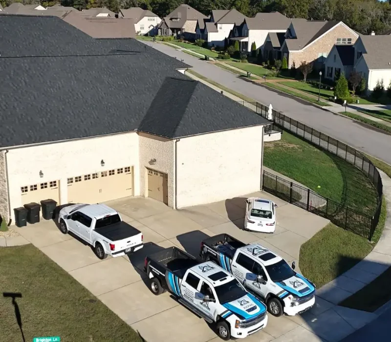 An aerial view of a large two-story residential home with a newly completed charcoal shingle roof, showing a concrete driveway where two branded J.L. Construction LLC work trucks and a white pickup are parked.